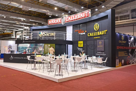 foto panoramica do stande da Barry Callebaut na FIPAN 2022 com dois andares, em sua maioria na cor preta com o logo de todas as marca. Na frente cadeiras e mesas brancas